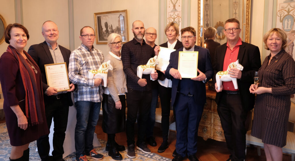 Personer står uppradade. Några håller i diplom och dalahästar.